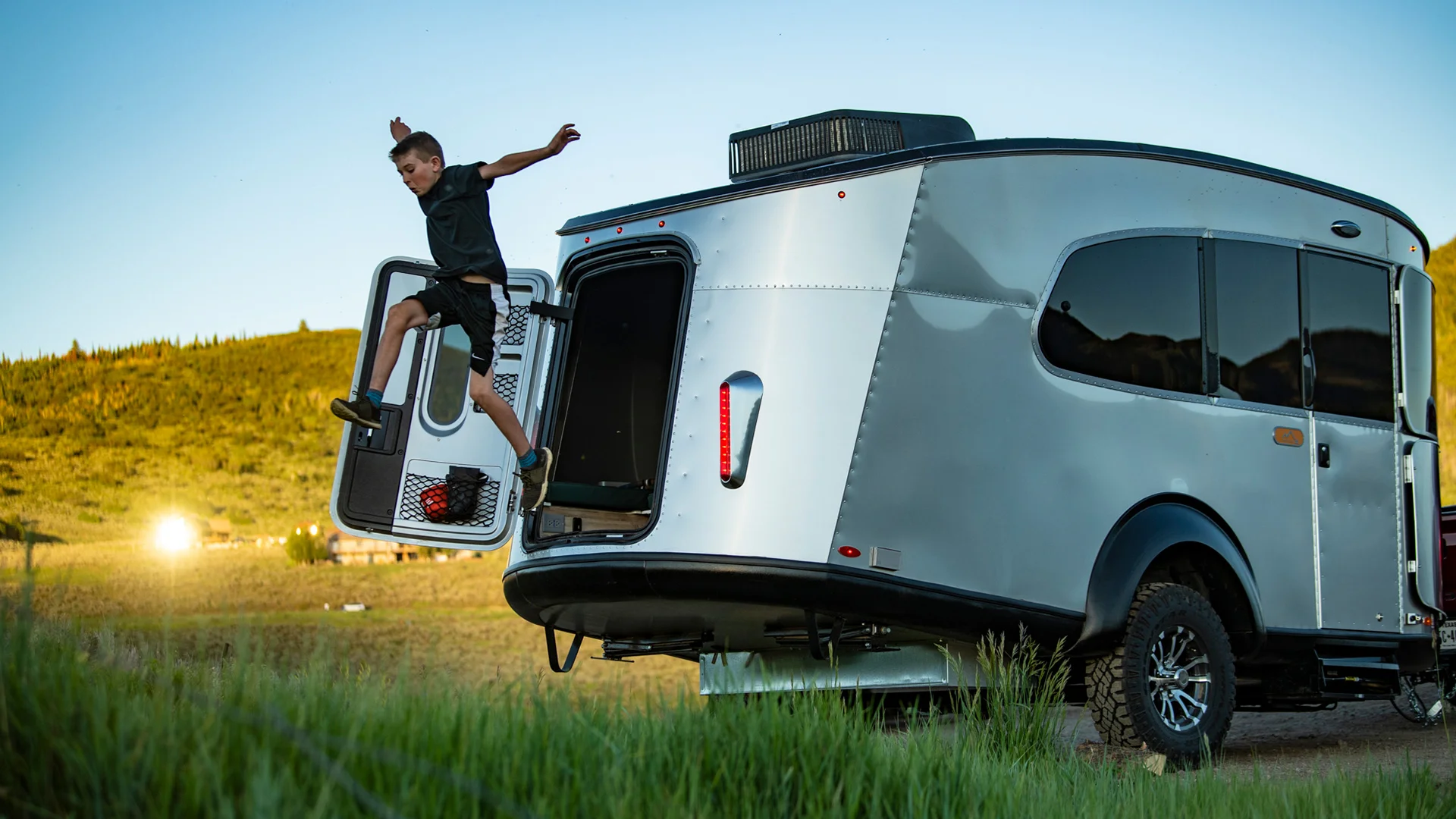 Boy jumping out from the open back of a modern silver camper van parked in a grassy field with a scenic view of hills and blue sky.