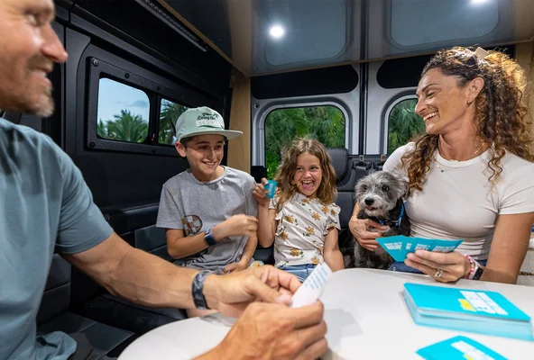 A family, including two children and a dog, happily play a card game inside a cozy van, surrounded by a lush green backdrop.