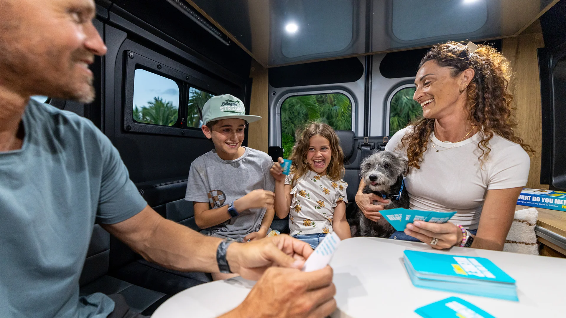 A family, including two children and a dog, happily play a card game inside a cozy van, surrounded by a lush green backdrop.