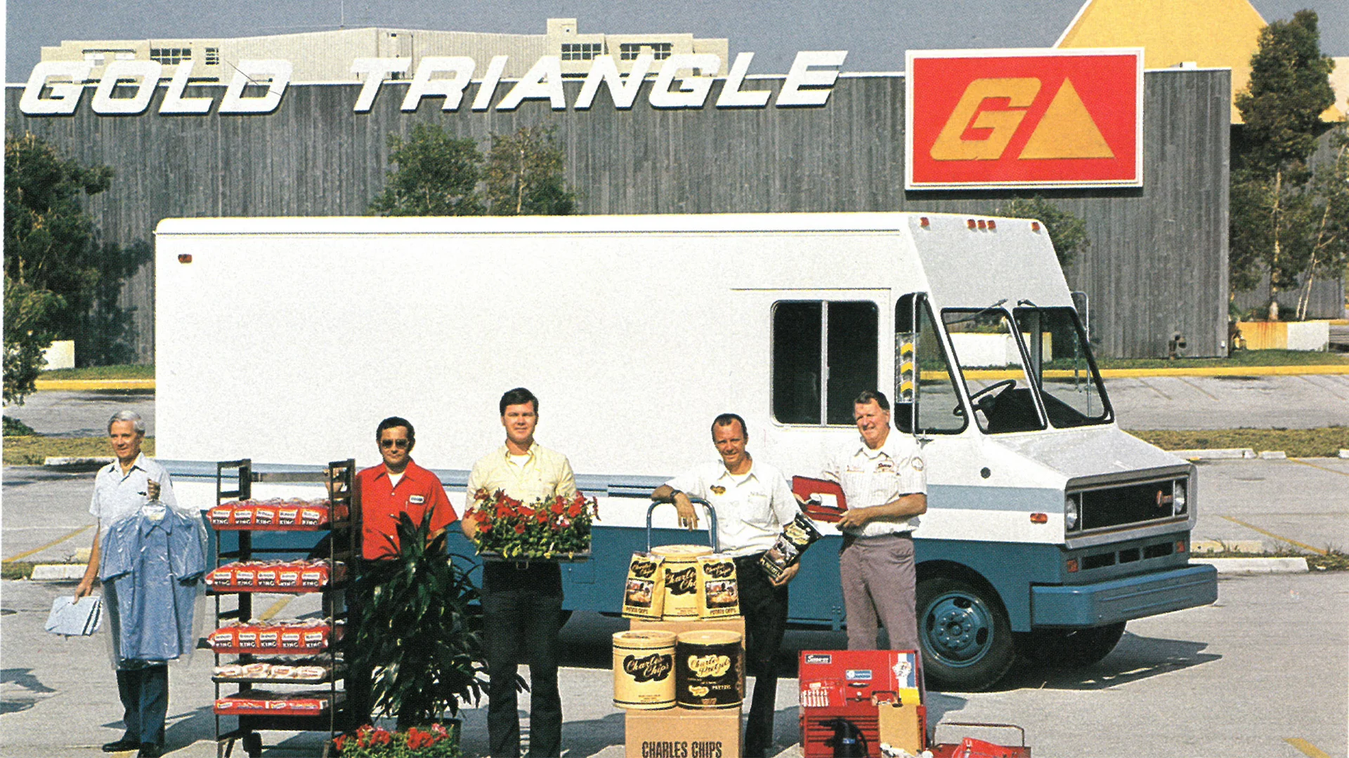 Five men stand in front of a delivery truck with various products, including plants and buckets, in a parking lot under "Gold Triangle" signage.