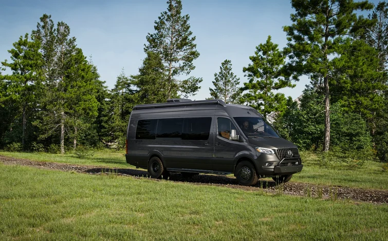 A black van parked on a gravel path surrounded by lush green grass and tall trees under a clear blue sky.
