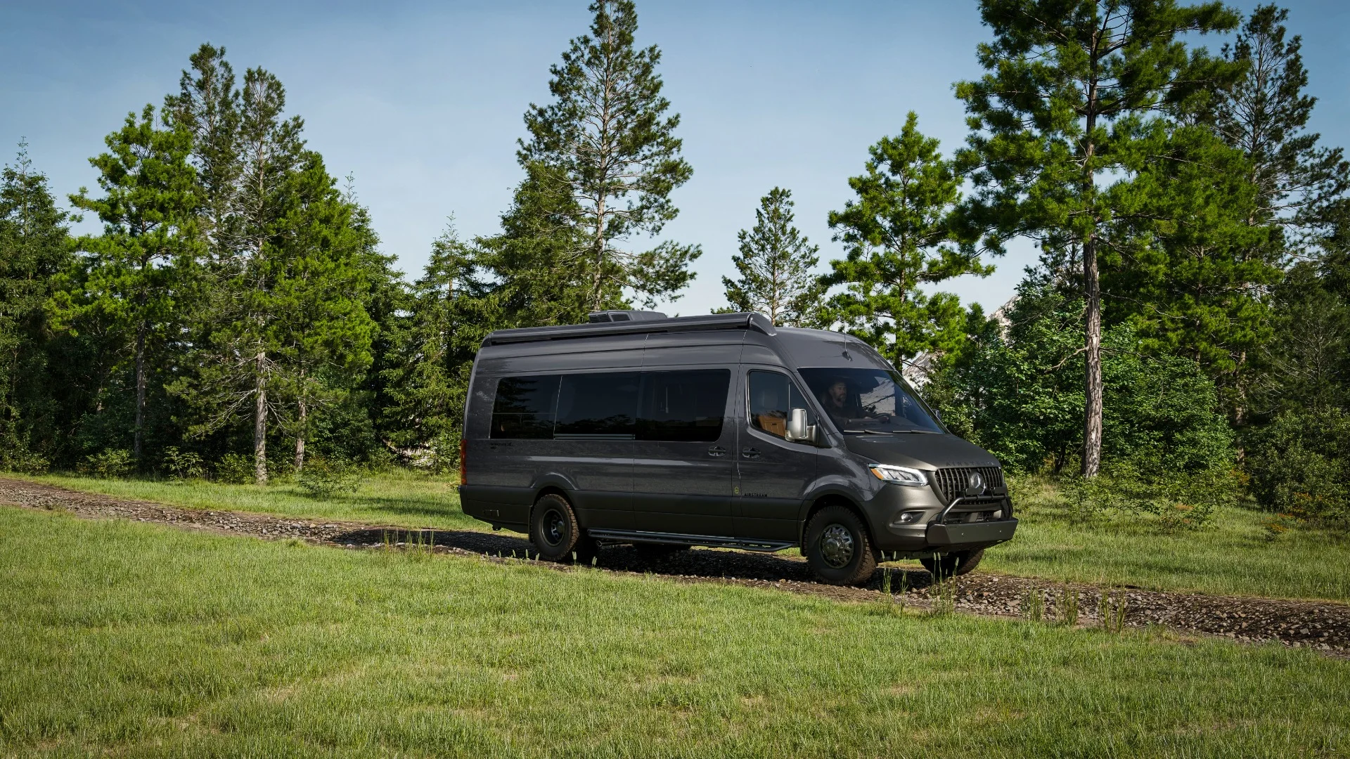 A black van parked on a gravel path surrounded by lush green grass and tall trees under a clear blue sky.