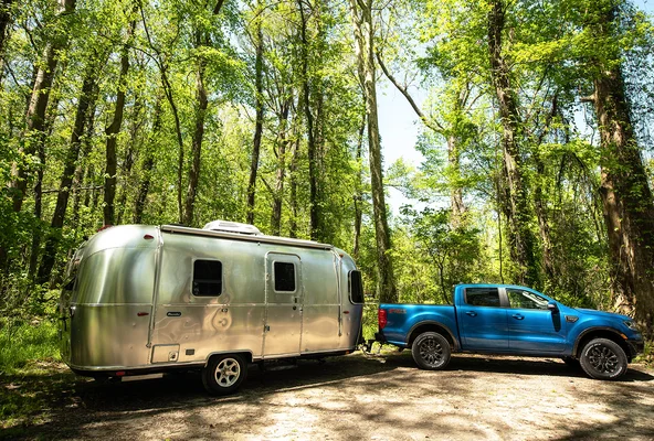 A blue pickup truck towing a silver Airstream trailer is parked in a lush forest with tall green trees under a clear sky.
