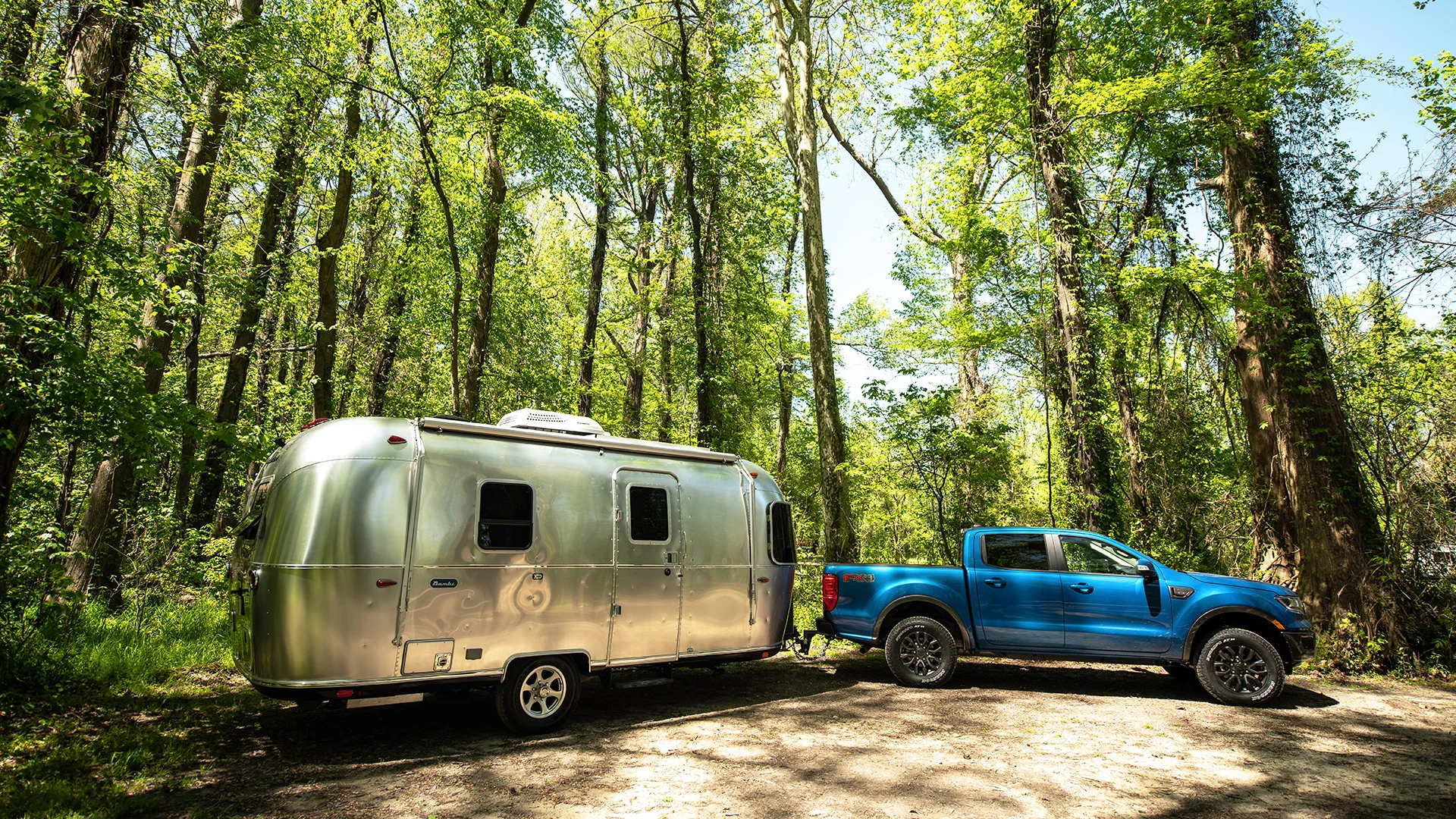 A blue pickup truck towing a silver Airstream trailer is parked in a lush forest with tall green trees under a clear sky.
