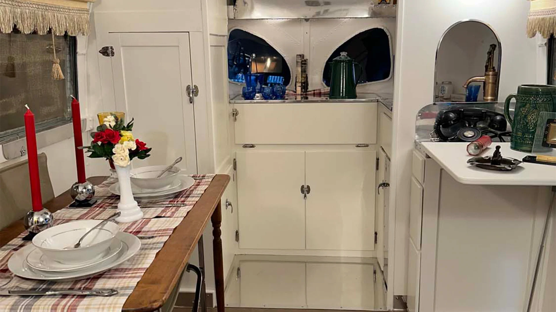 Compact RV interior with a dining table set for two, featuring red candles and flowers. Kitchen area includes a stove, sink, and green kettle.