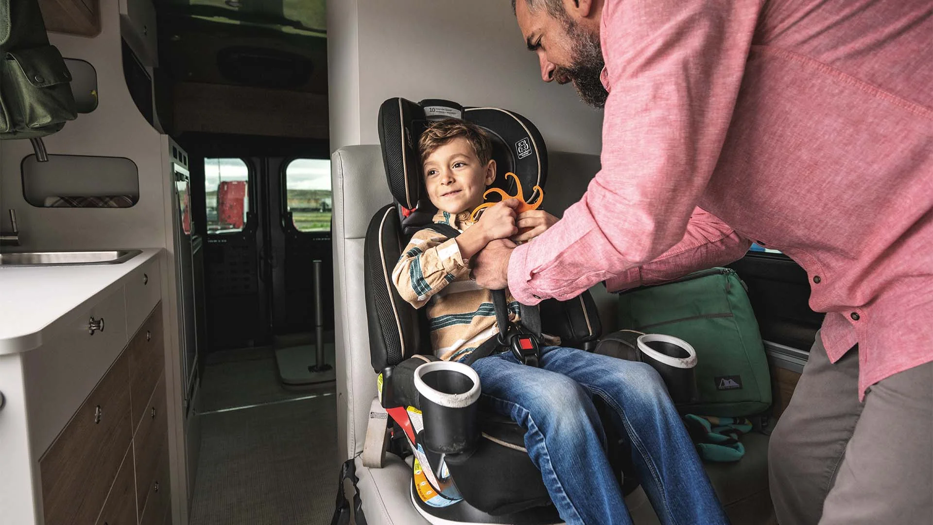 A child smiles in a car seat inside a van, while an adult in a pink shirt helps secure the seatbelt.
