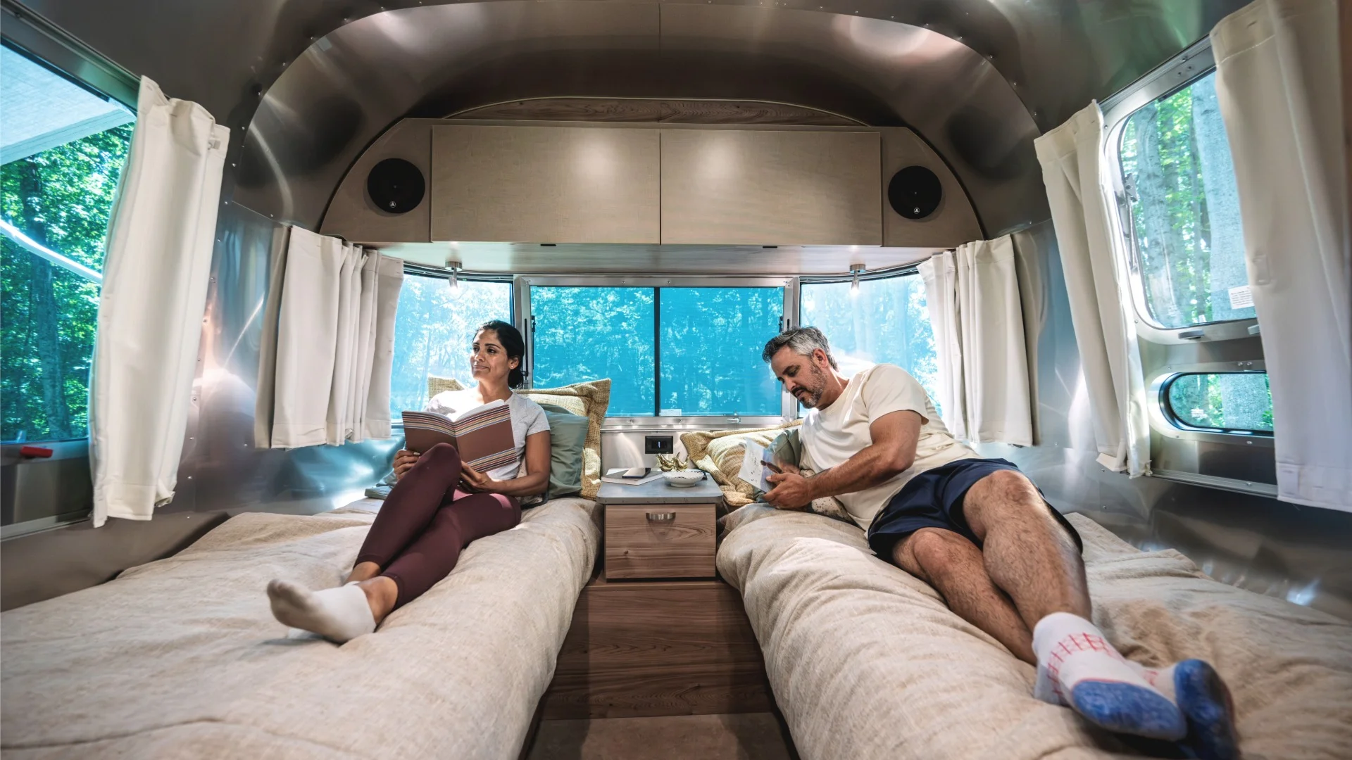 A couple relaxes in a trailer, both reading books, lying on separate beds with light streaming in through windows.