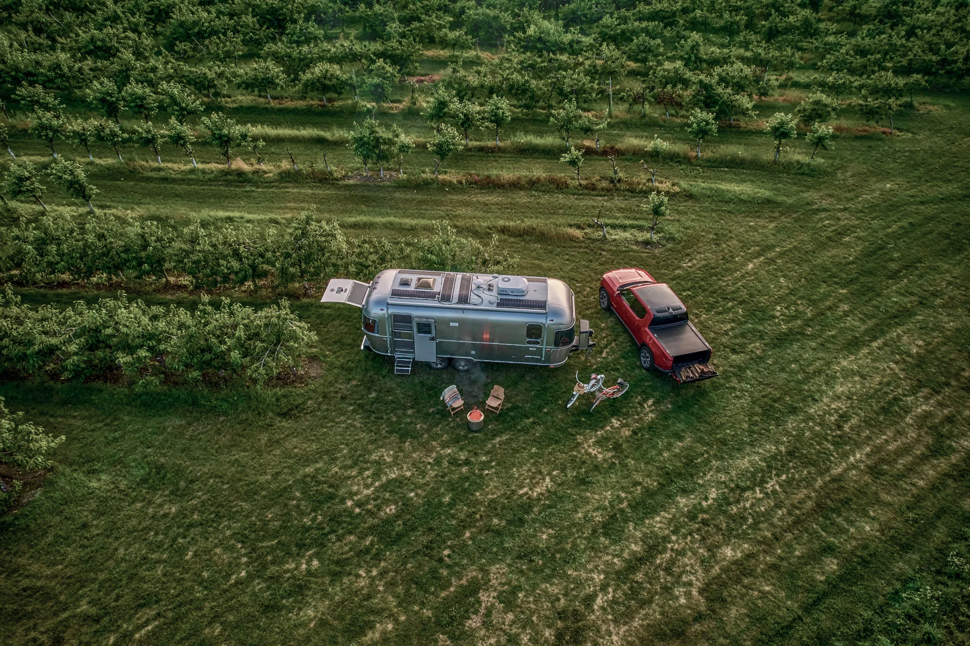 Aerial view of an RV and red pickup truck parked on grass near a vineyard, with chairs, a table, and a campfire setup beside them.