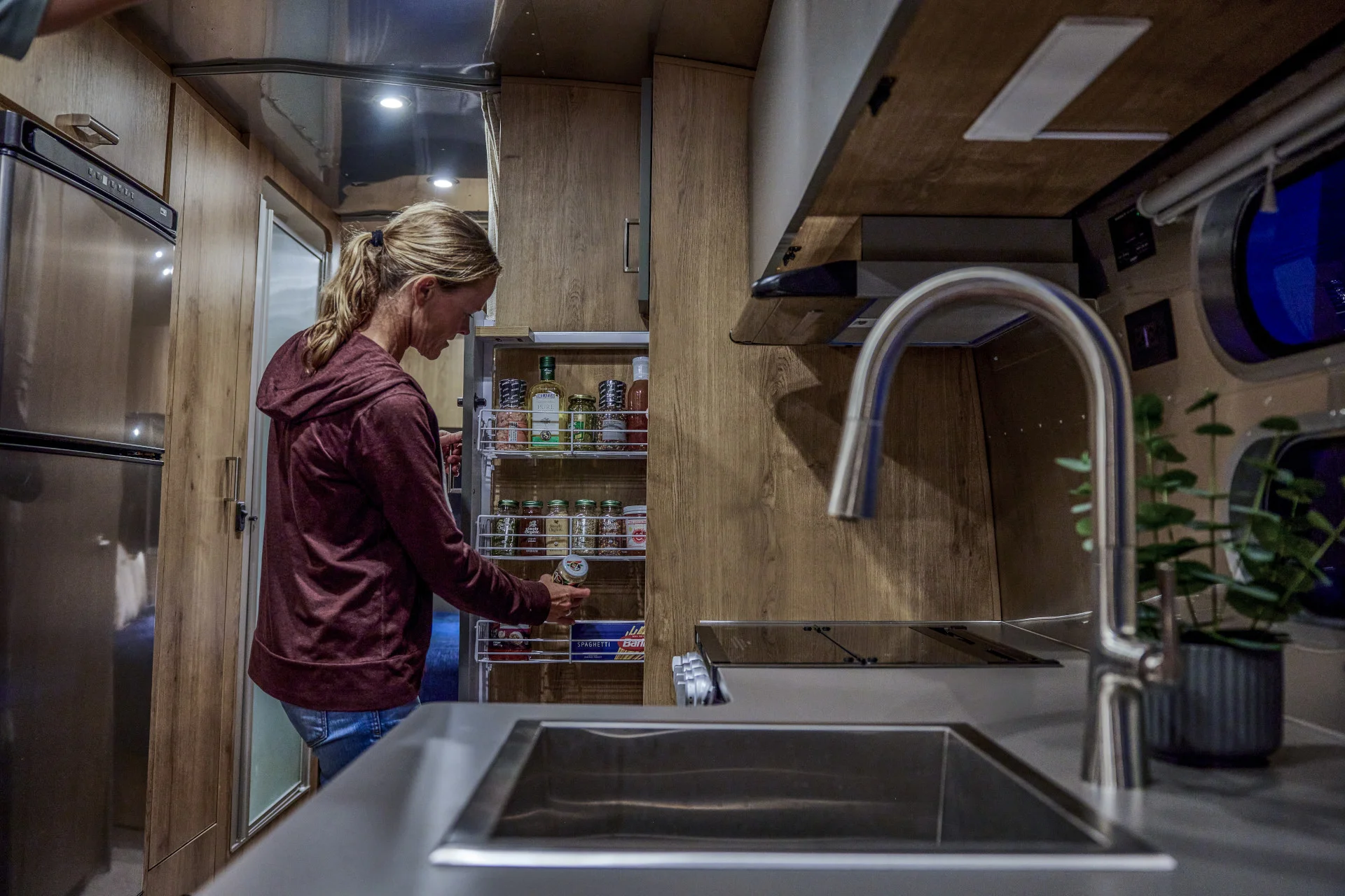 A person stands in a modern RV kitchen, reaching for items on a pull-out pantry shelf. Stainless steel appliances, wooden cabinets, and a sink with a tall faucet are visible.