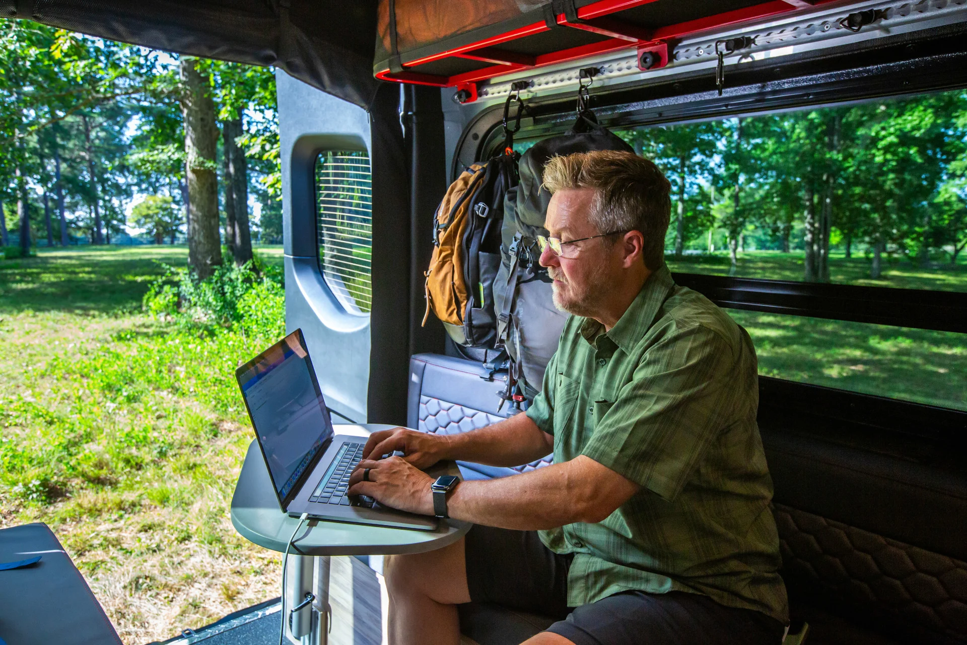 Man working on a laptop inside a camper van parked in a wooded area, wearing a green shirt and shorts, with a backpack hanging nearby.