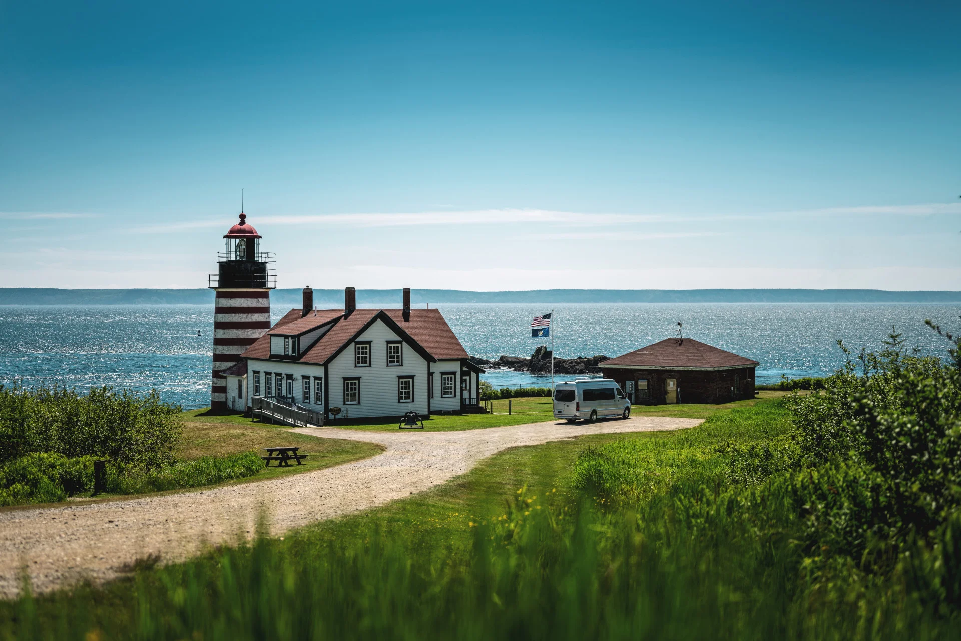 Lighthouse with a red roof near the sea, surrounded by greenery and a gravel path, with a parked van nearby under a clear blue sky.