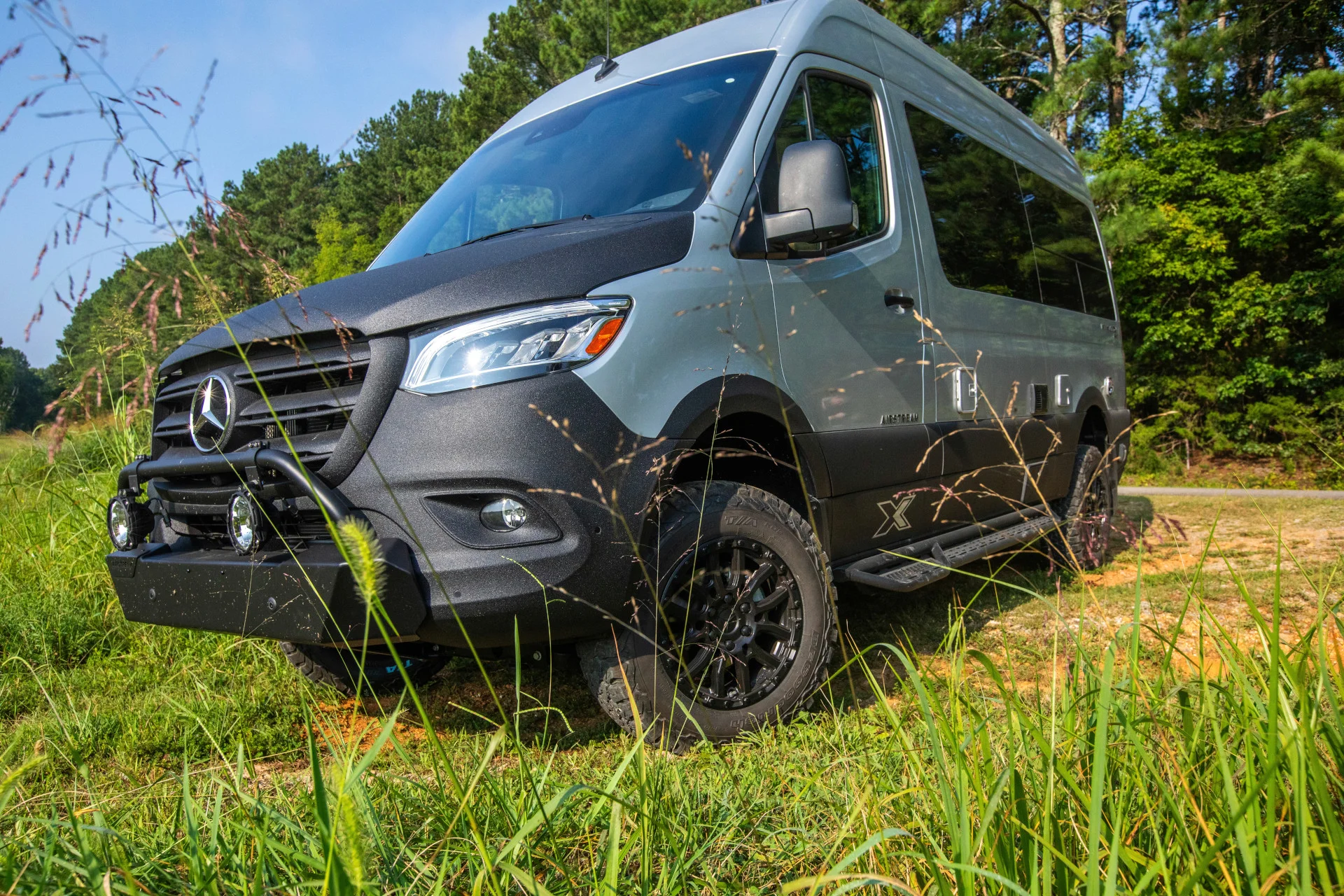 Off-road Mercedes-Benz van with rugged tires, parked on a grassy path, surrounded by tall grass and trees under a clear blue sky.