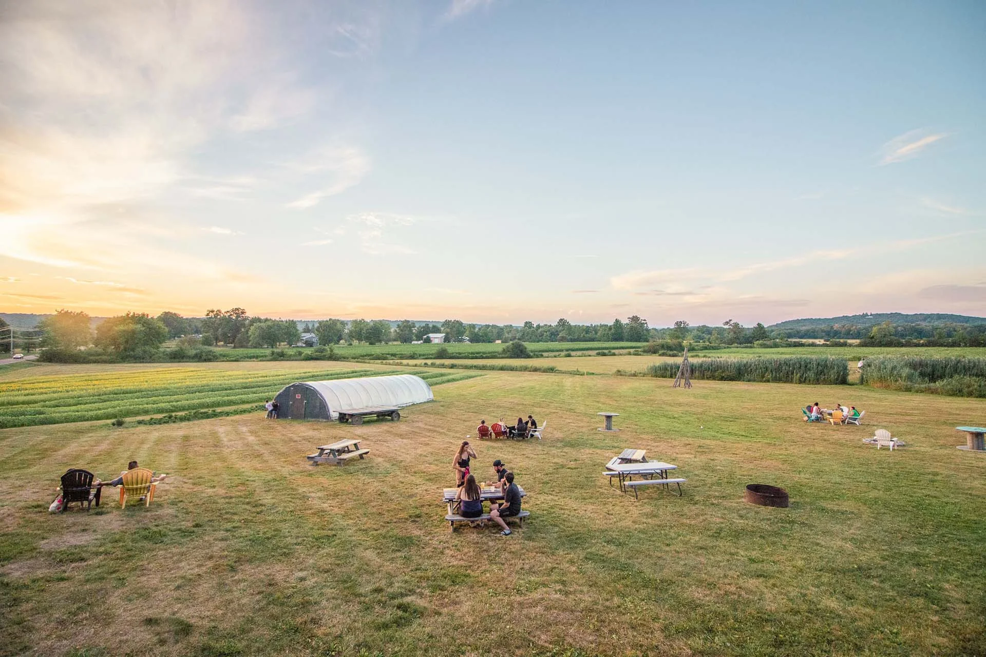 People gathered on a grassy field with tables and a distant view of a sunset over farmland.