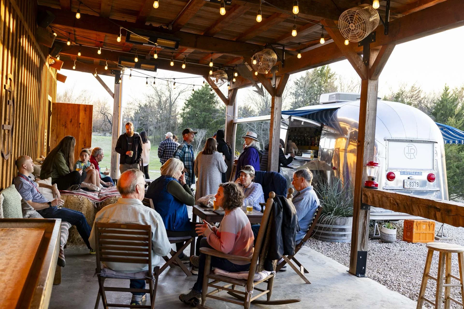 People gathered at an outdoor patio with string lights, sitting on wooden chairs, engaging in conversation near a vintage trailer.