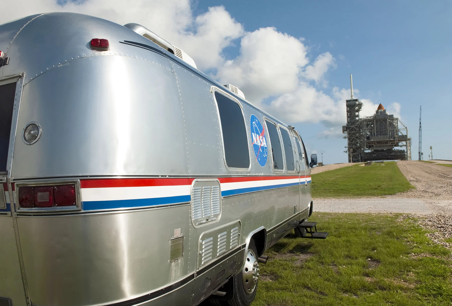 A shiny silver NASA vehicle, known as the Astrovan, is parked on grassy ground with a space shuttle on a launch pad visible in the background under a partly cloudy sky.