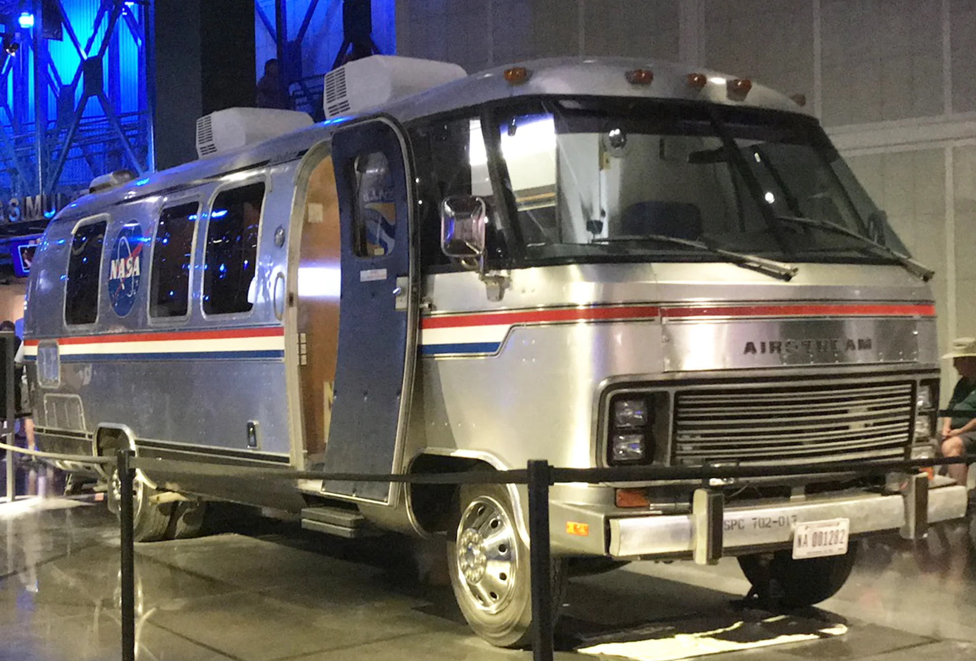 A vintage silver Airstream motorhome with NASA markings and a red, white, and blue stripe is displayed indoors behind a rope barrier, with its door open and people nearby.