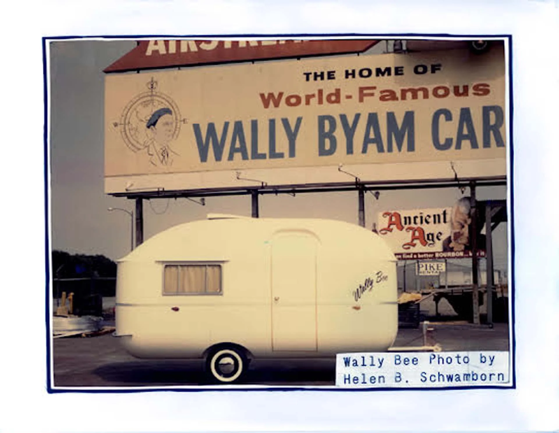 A small vintage trailer with "Wally Bee" on its side is parked in front of a "World-Famous Wally Byam" billboard.