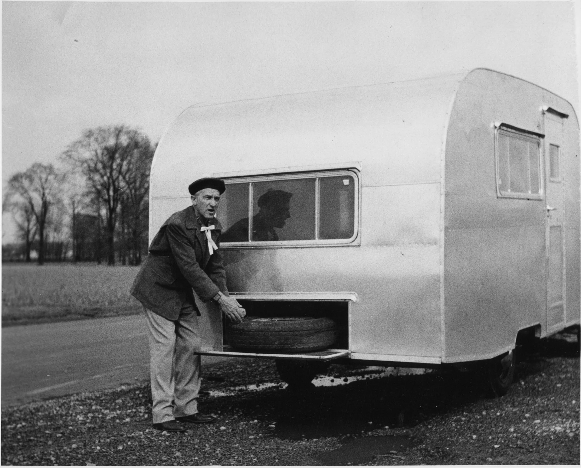 An older man wearing a beret and scarf loads or unloads a spare tire from the front storage compartment of a silver travel trailer parked by a rural road. Trees and fields are visible in the background.