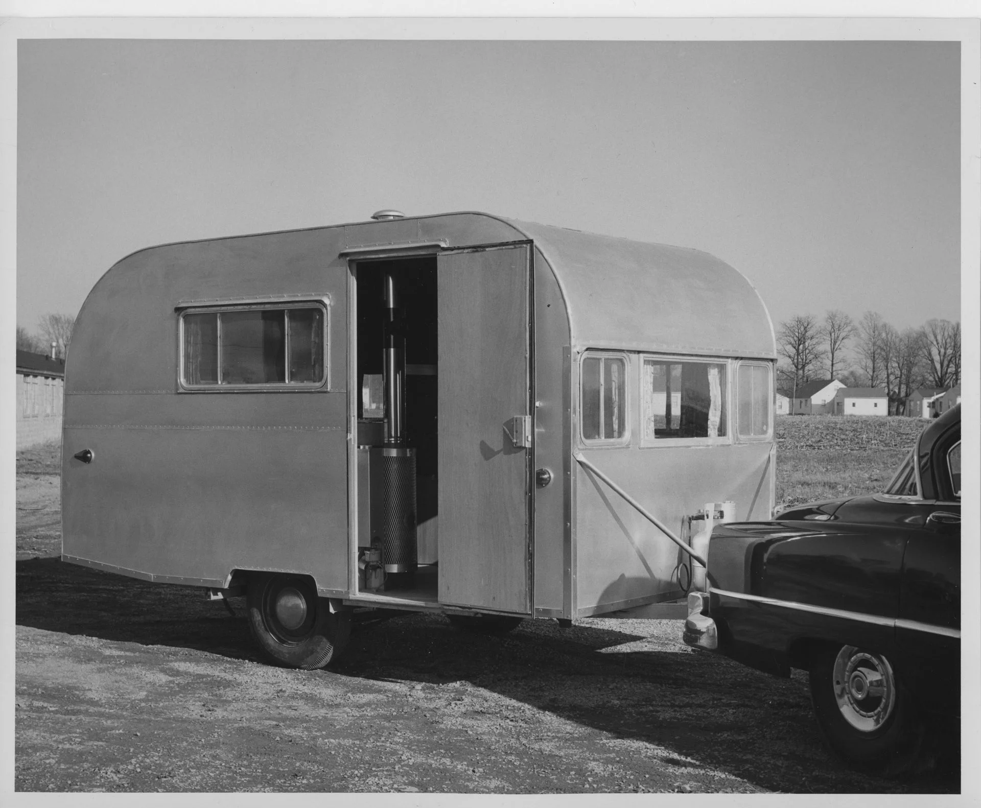 A vintage travel trailer hitched to a car is parked on a gravel lot. The trailer door is open, revealing part of the interior. Leafless trees and small buildings are visible in the background.