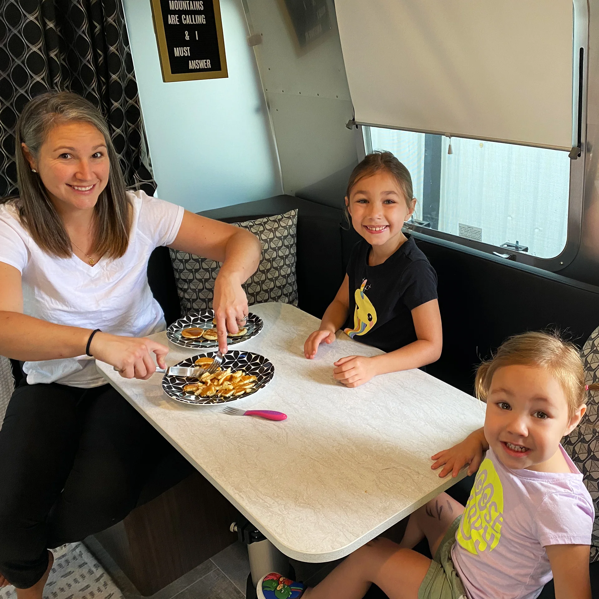 A woman and two girls sitting at a table in a camper, smiling, with plates of food in front of them.