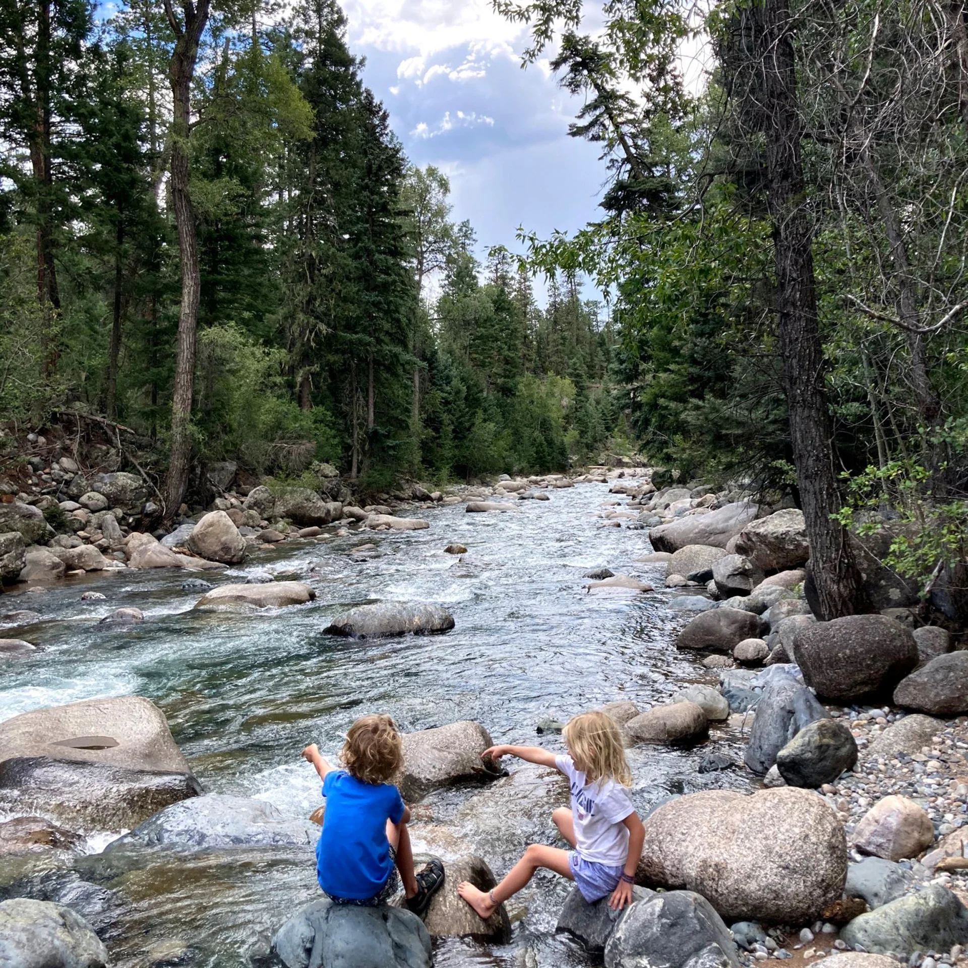 Two children sit on rocks by a flowing river in a forest. Trees line the riverbank under a partly cloudy sky.