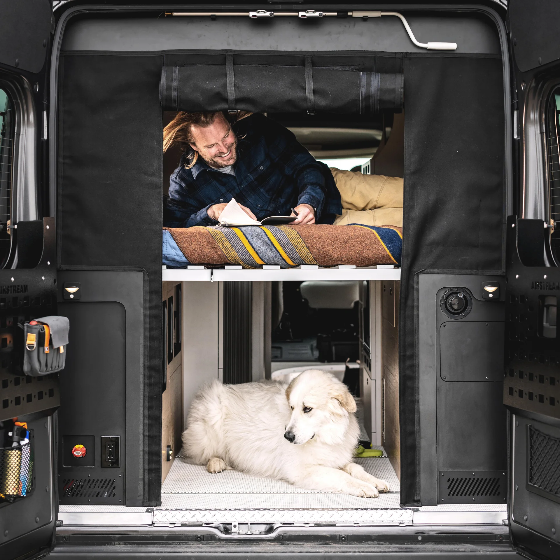 Person writing in a cozy camper van with open doors, a white dog resting below.