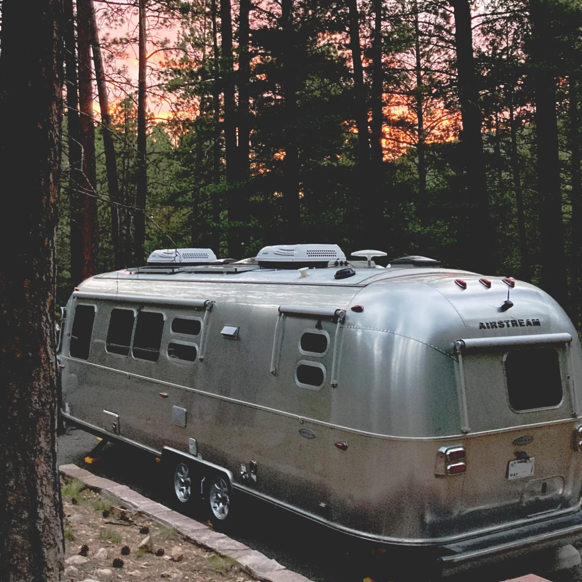 A silver Airstream trailer is parked among tall pine trees at dusk, with a soft orange and pink sunset visible through the forest in the background.