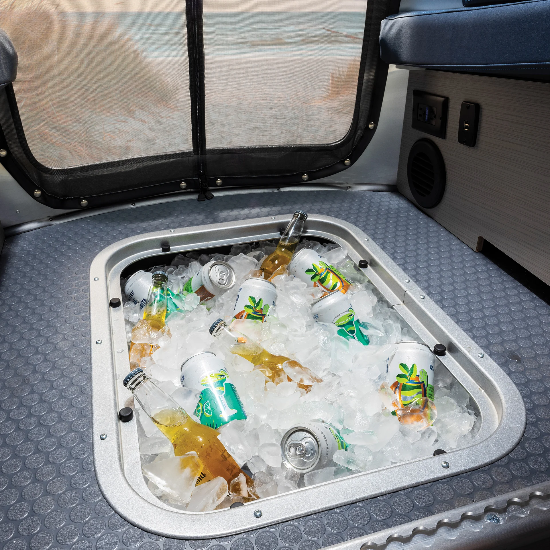 A cooler inside a vehicle is filled with ice, assorted beverage bottles, and cans. A sandy beach is visible through the open rear door.