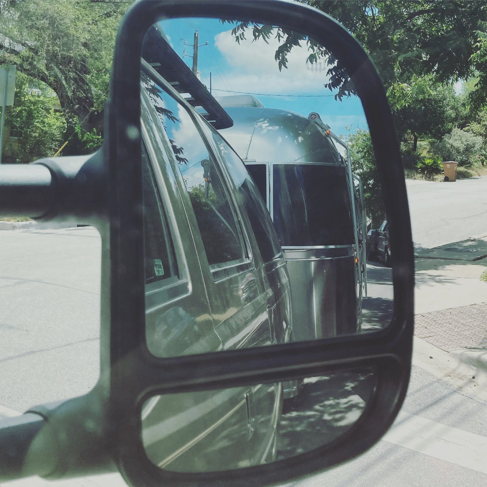 A cars side mirror reflects an Airstream trailer being towed along a tree-lined suburban street on a sunny day.