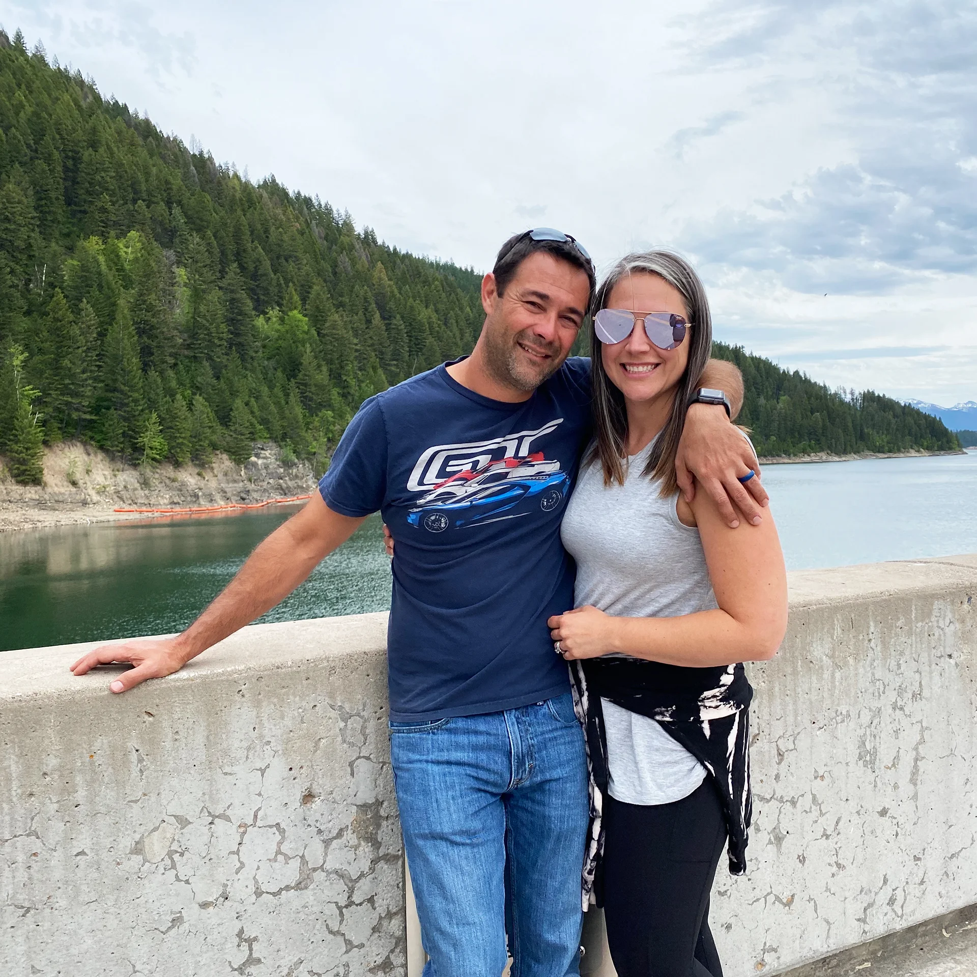 A couple stands smiling by a concrete barrier, with lush green trees and a calm lake in the background.
