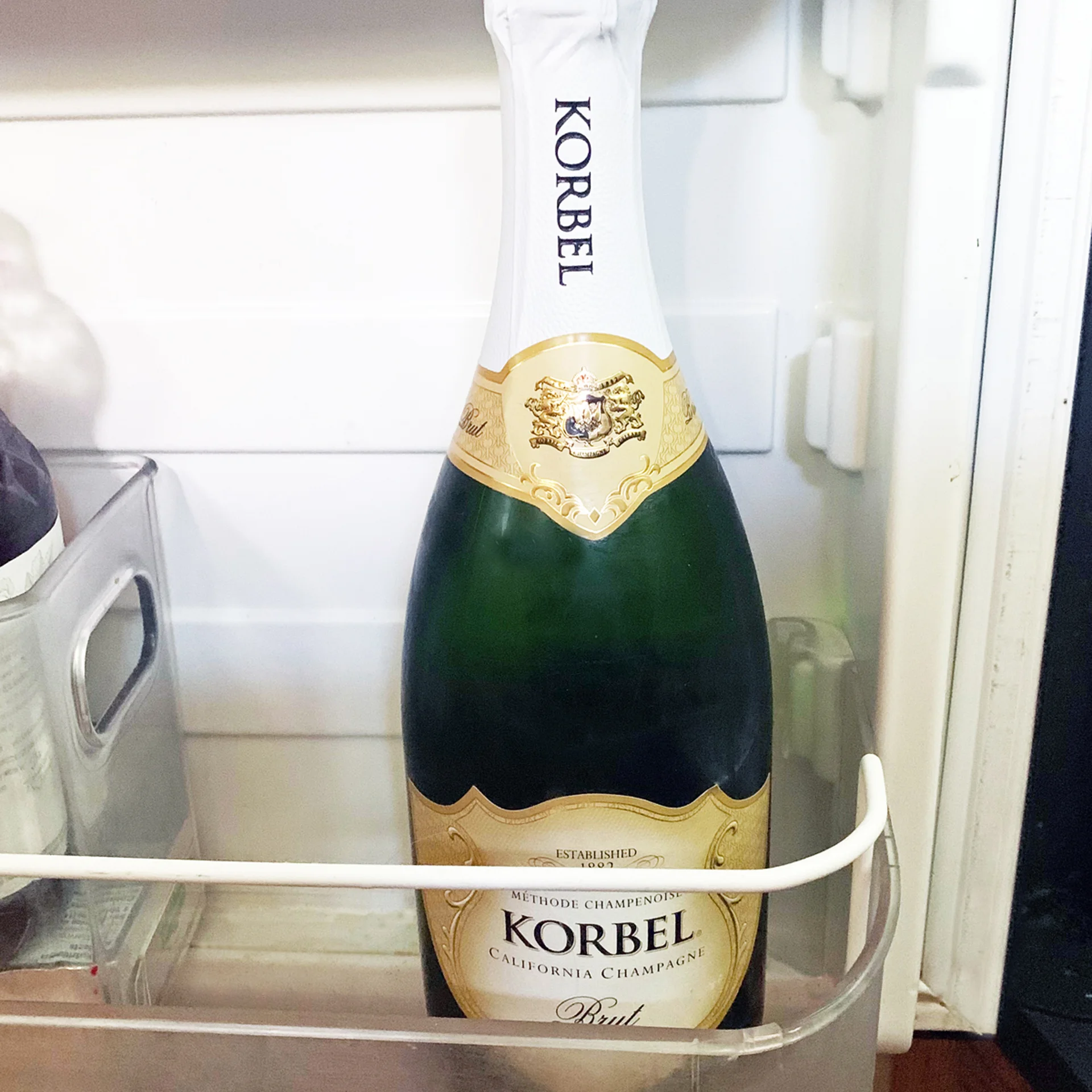 A bottle of Korbel California Champagne Brut is standing upright on a refrigerator door shelf, with white and gold labeling visible.