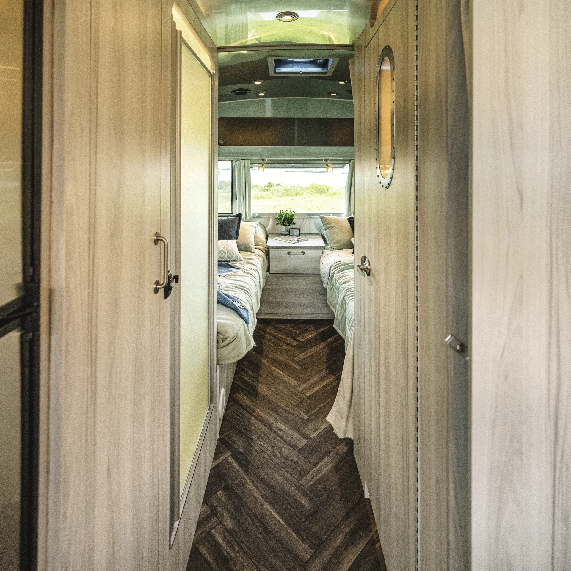 Interior view of a modern RV hallway with wood flooring, light wood walls, and a glimpse of a bedroom area at the end featuring two beds, a center drawer, a plant, and large windows letting in natural light.