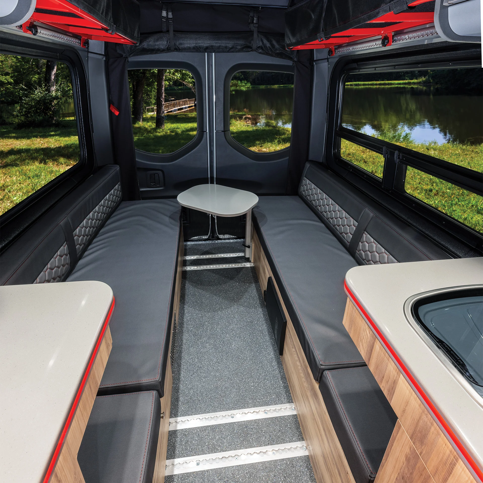 Interior of a camper van with two long cushioned benches facing each other, a small oval table in the center, large windows showing greenery and a pond, and part of a kitchenette visible on the right.