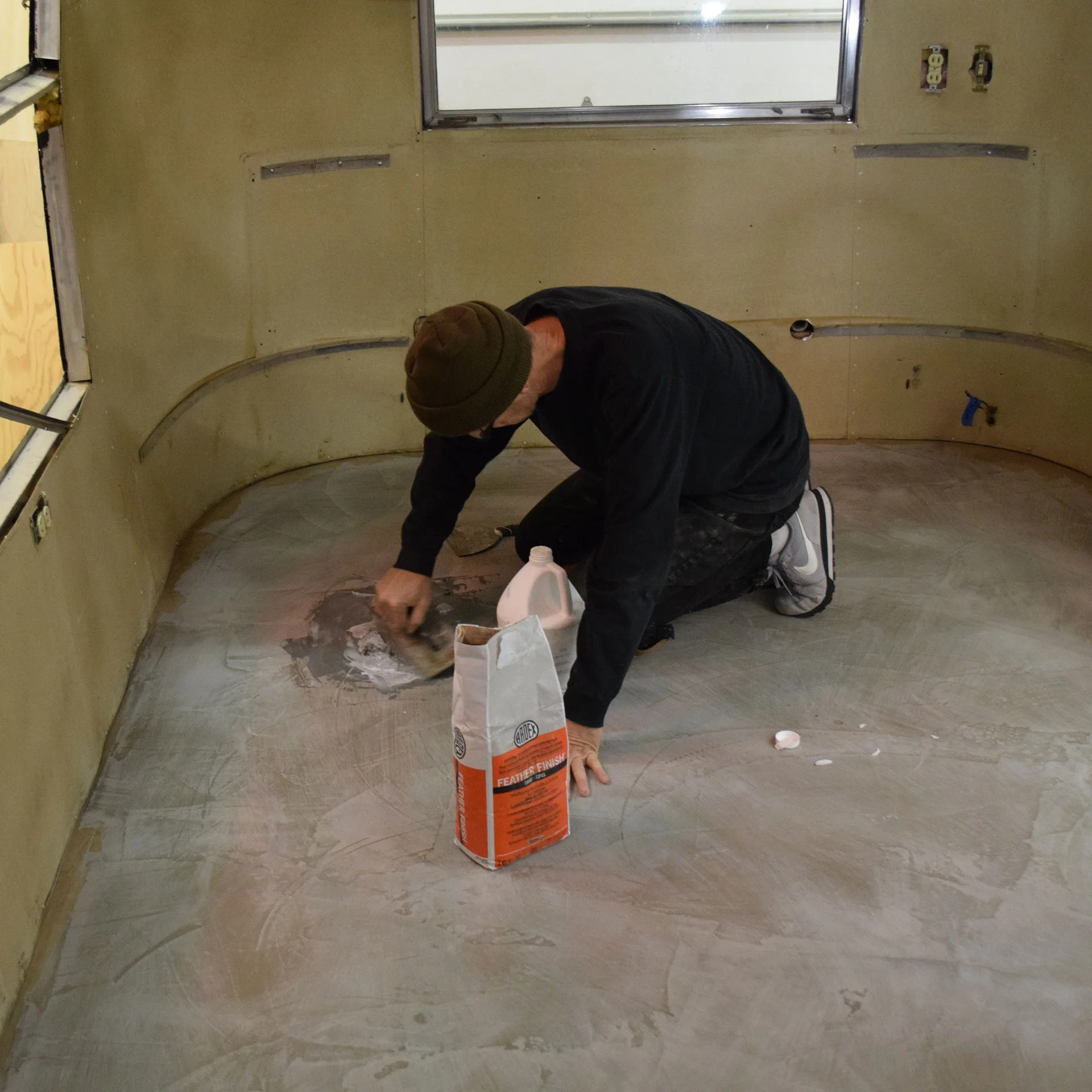 A person wearing a beanie and dark clothing kneels on the floor of a bare room, spreading a powdered material from an orange bag onto the floor with a trowel. The walls appear unfinished and there are windows in the room.
