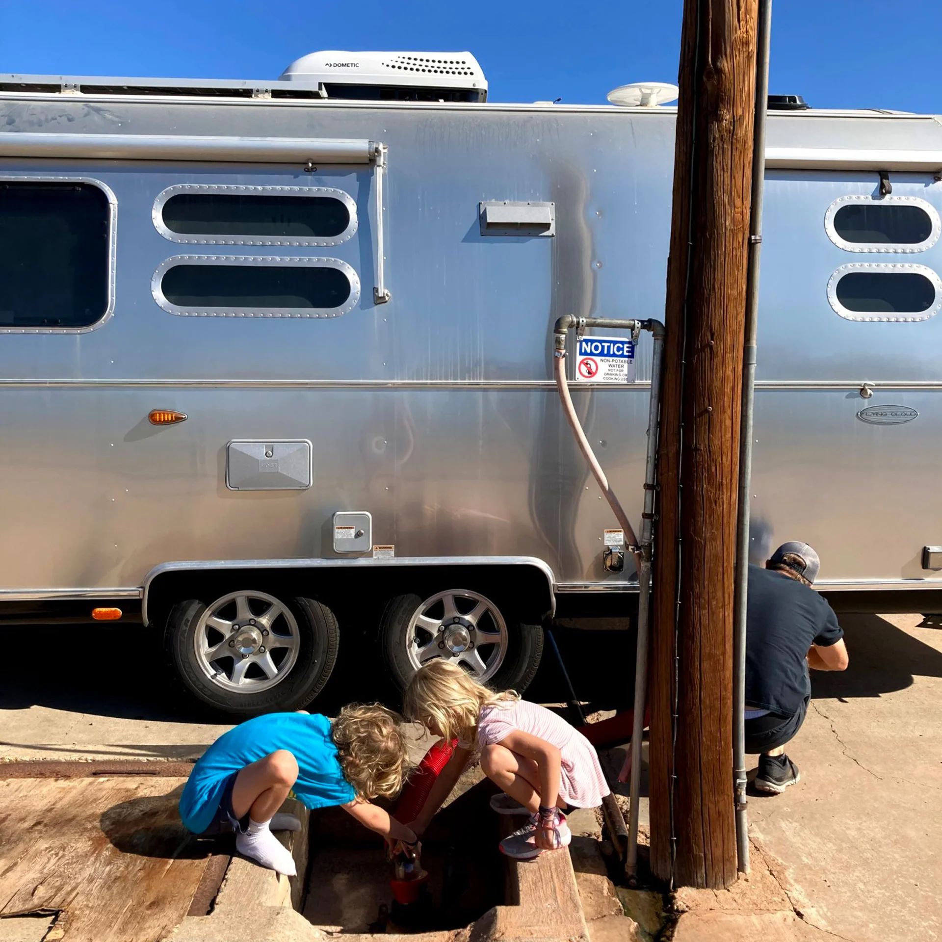 Two young children crouch next to a drain in front of a silver Airstream trailer, while an adult kneels by a wooden pole nearby, working on something out of view. Bright sunlight casts clear shadows on the scene.