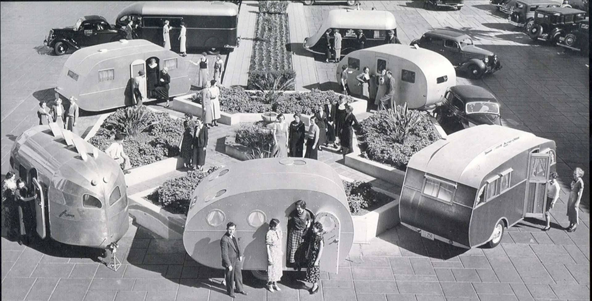 A vintage scene with people admiring various unique, futuristic trailers placed around a geometric garden in a plaza, with classic cars nearby.