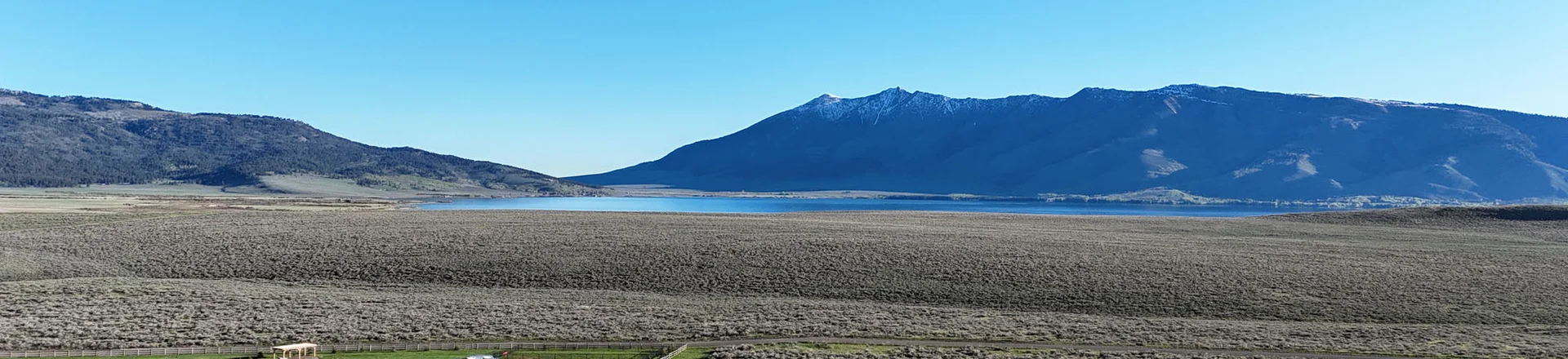 A campground with several RVs and vehicles is set in a green, grassy area surrounded by sagebrush, with a lake and snow-capped mountains under a clear blue sky in the background.