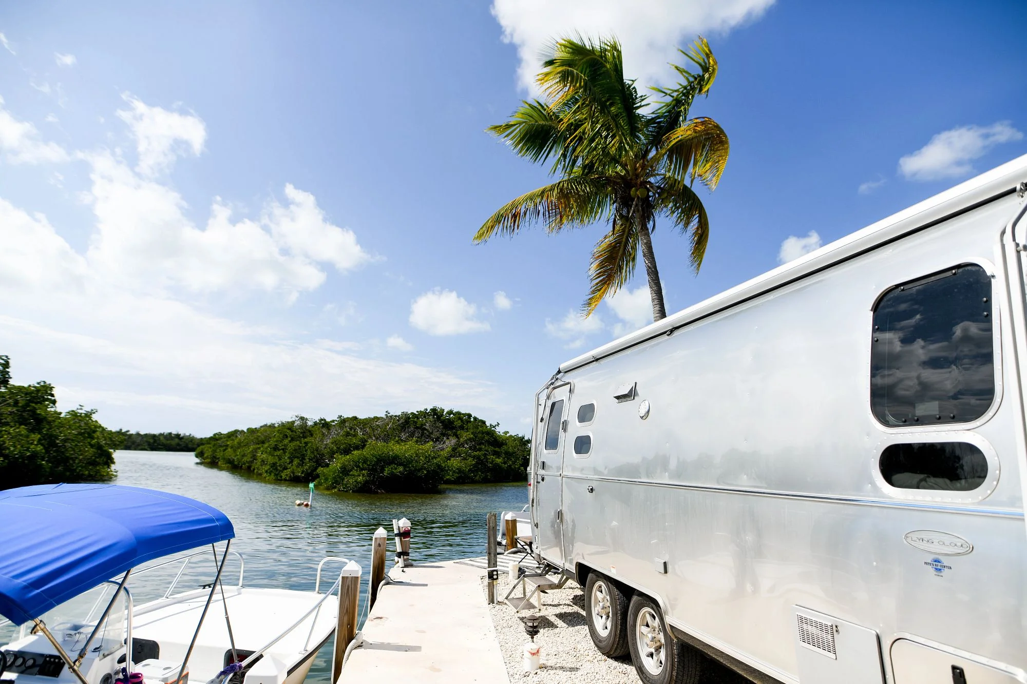 A silver trailer is parked by a scenic waterfront with a blue and white boat, lush greenery, and a palm tree under a clear blue sky.