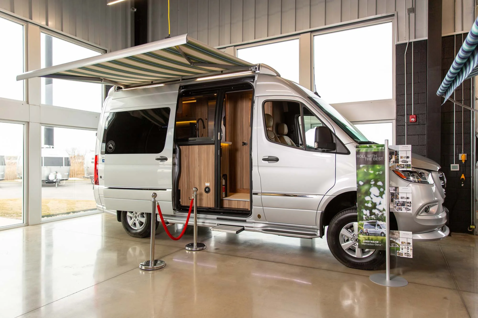 Silver camper van on display indoors, featuring an extended striped awning and open door, with informational signs nearby.
