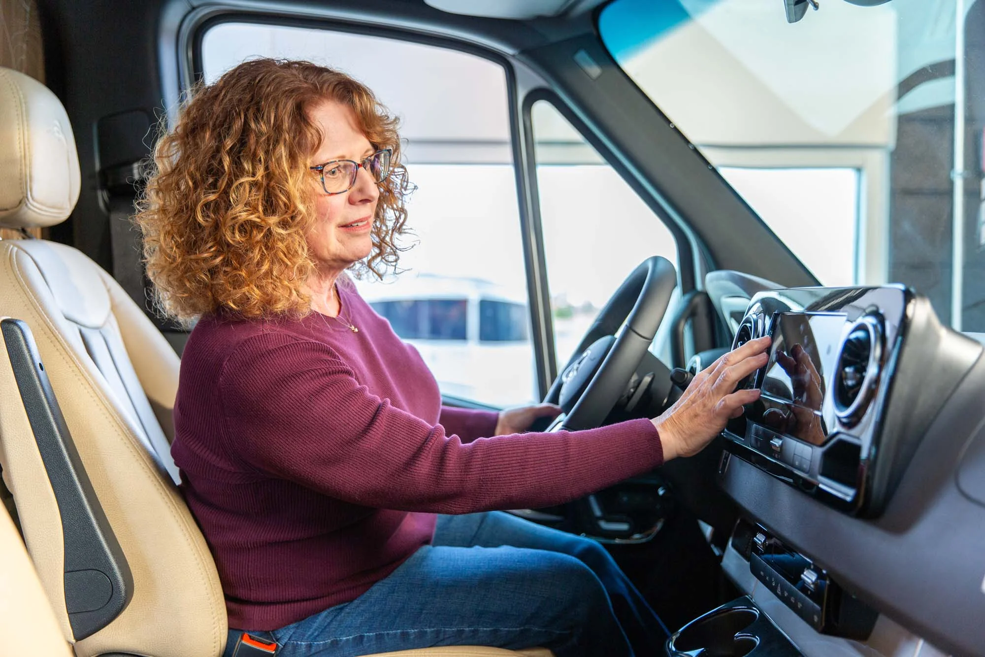 A person with curly hair and glasses sits in a vehicle's driver seat, adjusting the dashboard controls.