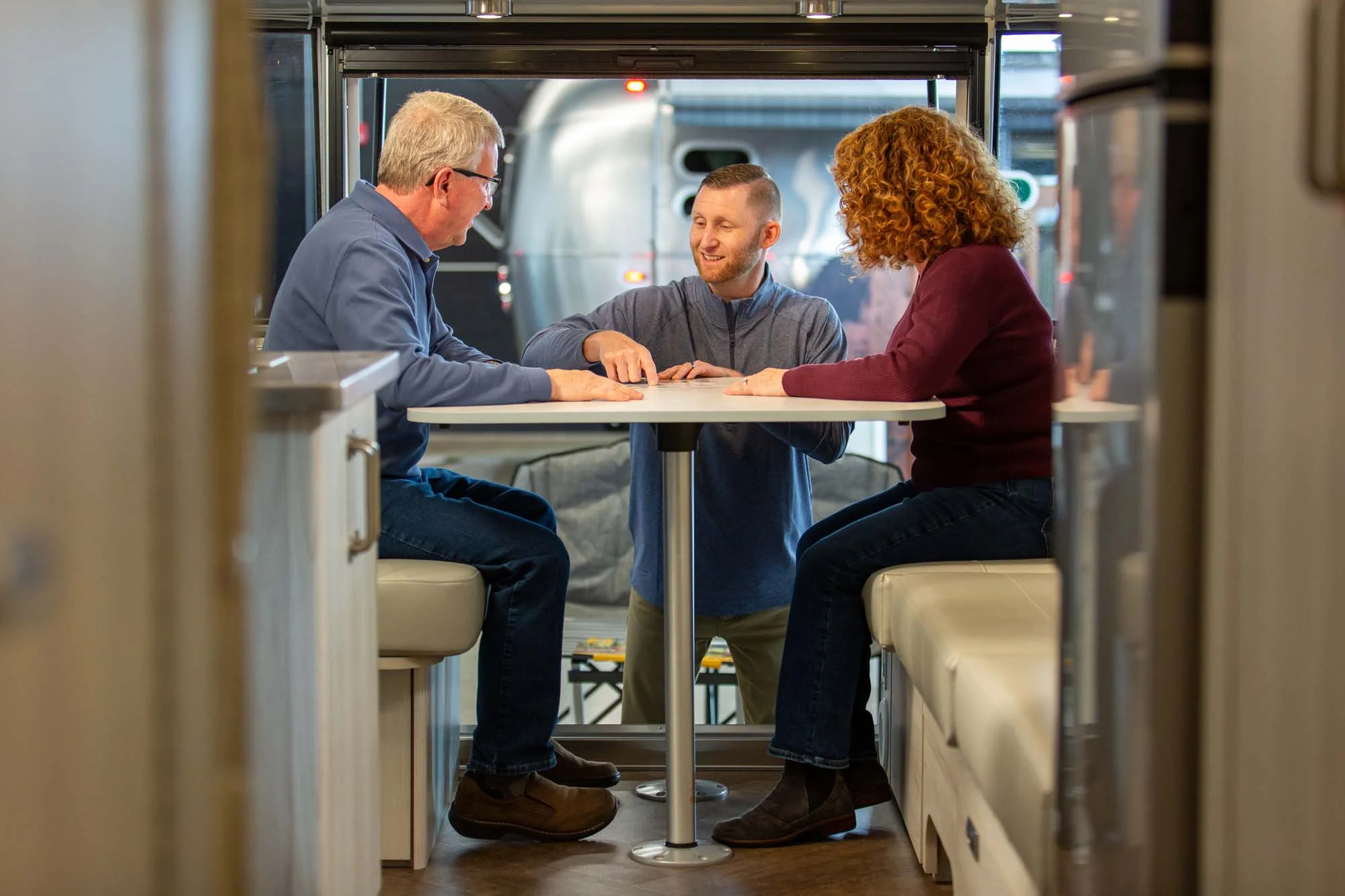 Three people sit at a table inside an RV, engaged in conversation. The interior is modern, with soft seating and a large window behind them.