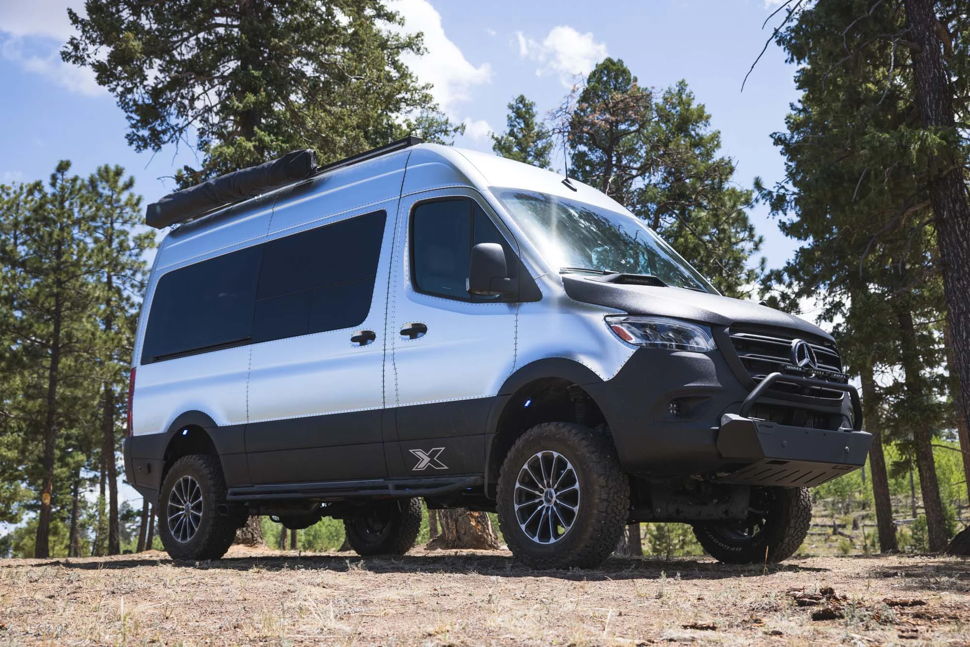 Silver and black off-road van parked in a forested area, surrounded by tall trees under a partly cloudy sky.