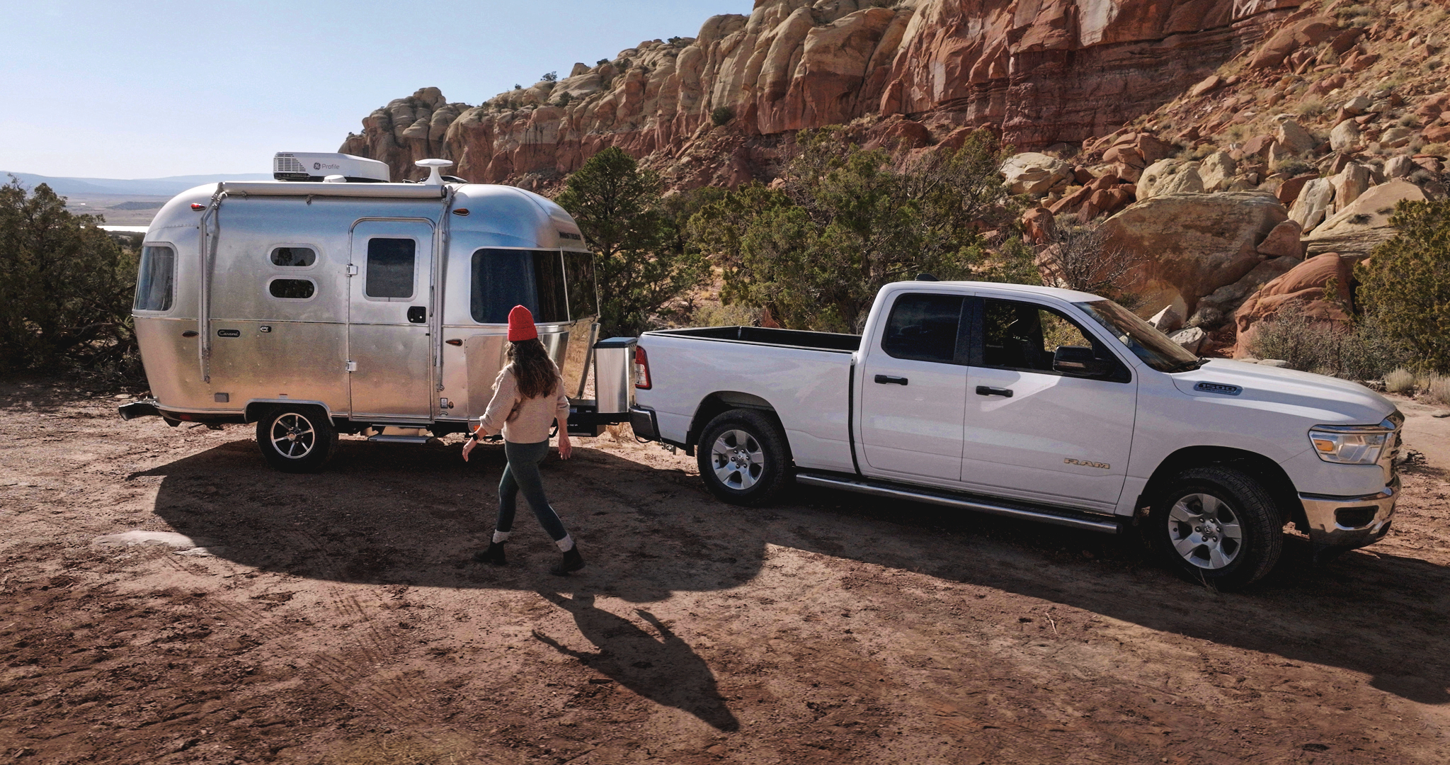 A person wearing a red beanie walks towards a silver Airstream trailer hitched to a white pickup truck in a rocky desert landscape.