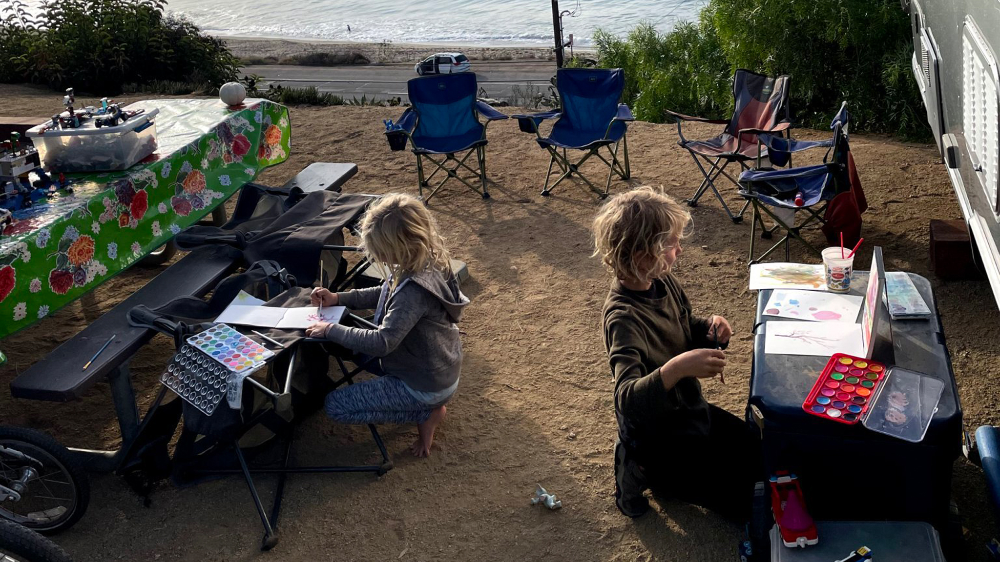 Two children create watercolor paintings at a campsite near the beach. Art supplies and picnic tables are scattered around, and empty camping chairs face the ocean in the background.