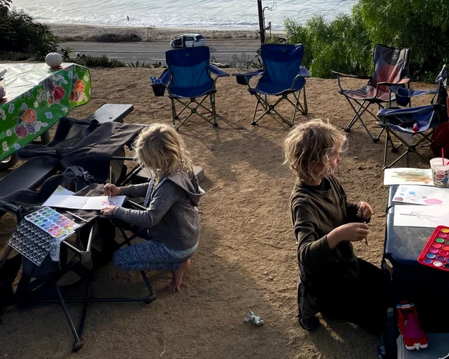 Two kids sitting outside of their Airstream travel trailer at their own tables while they paint next to the water.|Three kids playing in the ocean.|Four kids sitting on the edge of a boat with lifejackets on looking out at the water.|A young girl in a blue jacket carrying a bucket of things she found as she walks along a beach.|A young kid sitting at a picnic table writing about nature|Two kids sitting at a table outside of their Airstream travel trailer while doing crafts.|A young kid writes in a notebook about what they found in nature.|A father and two kids sitting outside of their Airstream travel trailer while they learn and write about their adventures.|Two kids sitting in the galley of their Airstream travel trailer while they learn on the road.|Two kids sitting outside of their Airstream travel trailer at their own tables while they paint next to the water.