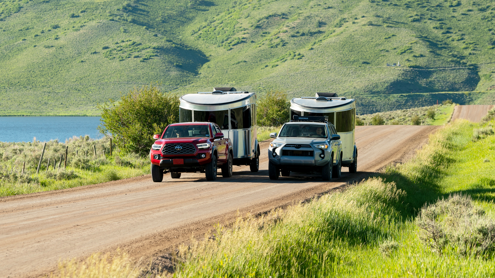 Two trucks towing trailers on a scenic dirt road beside a lake and green hills under a clear sky.