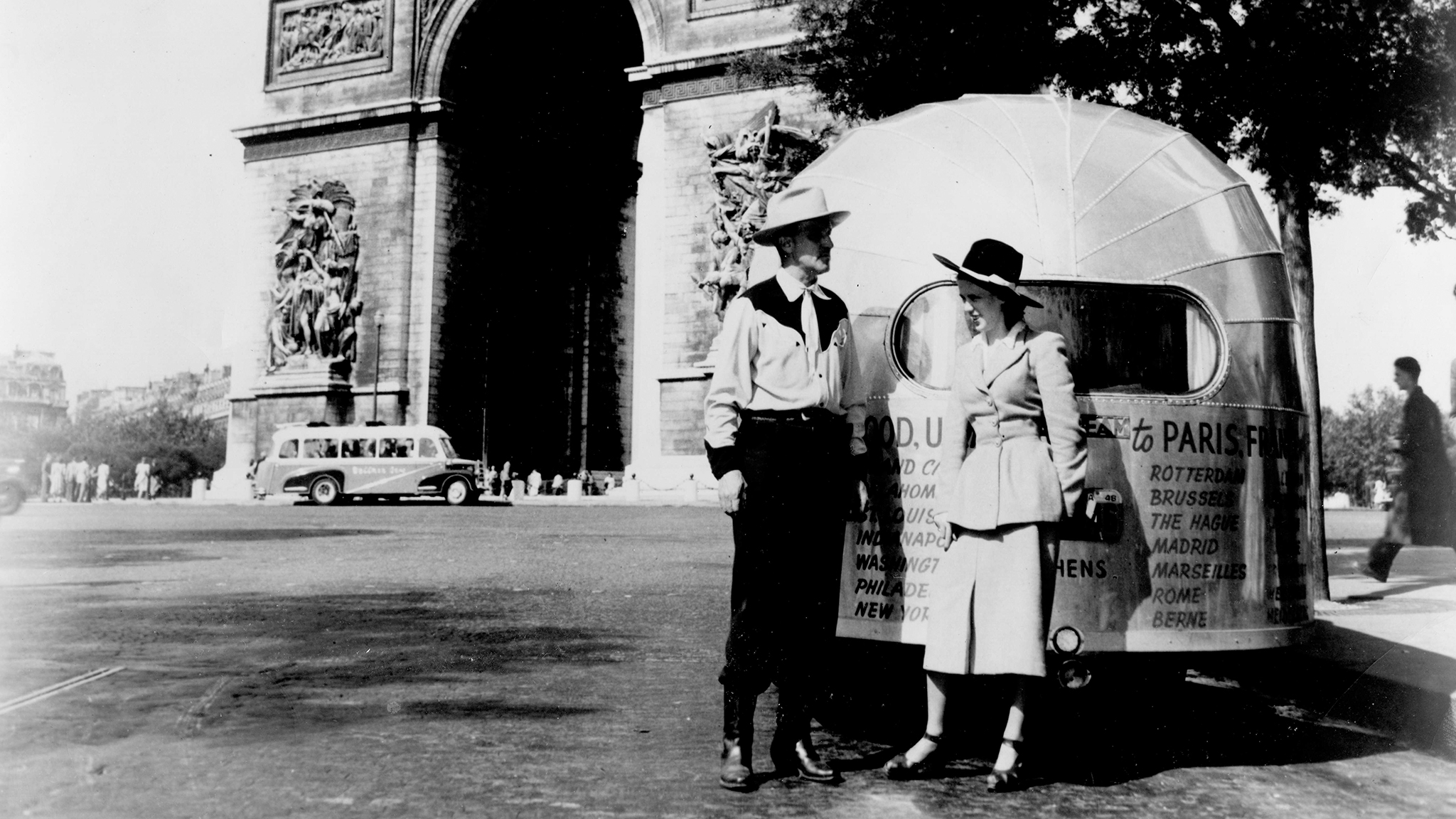 A black-and-white photo shows two people in vintage clothing standing beside an old trailer in front of the Arc de Triomphe in Paris. An early automobile and several pedestrians are visible in the background.