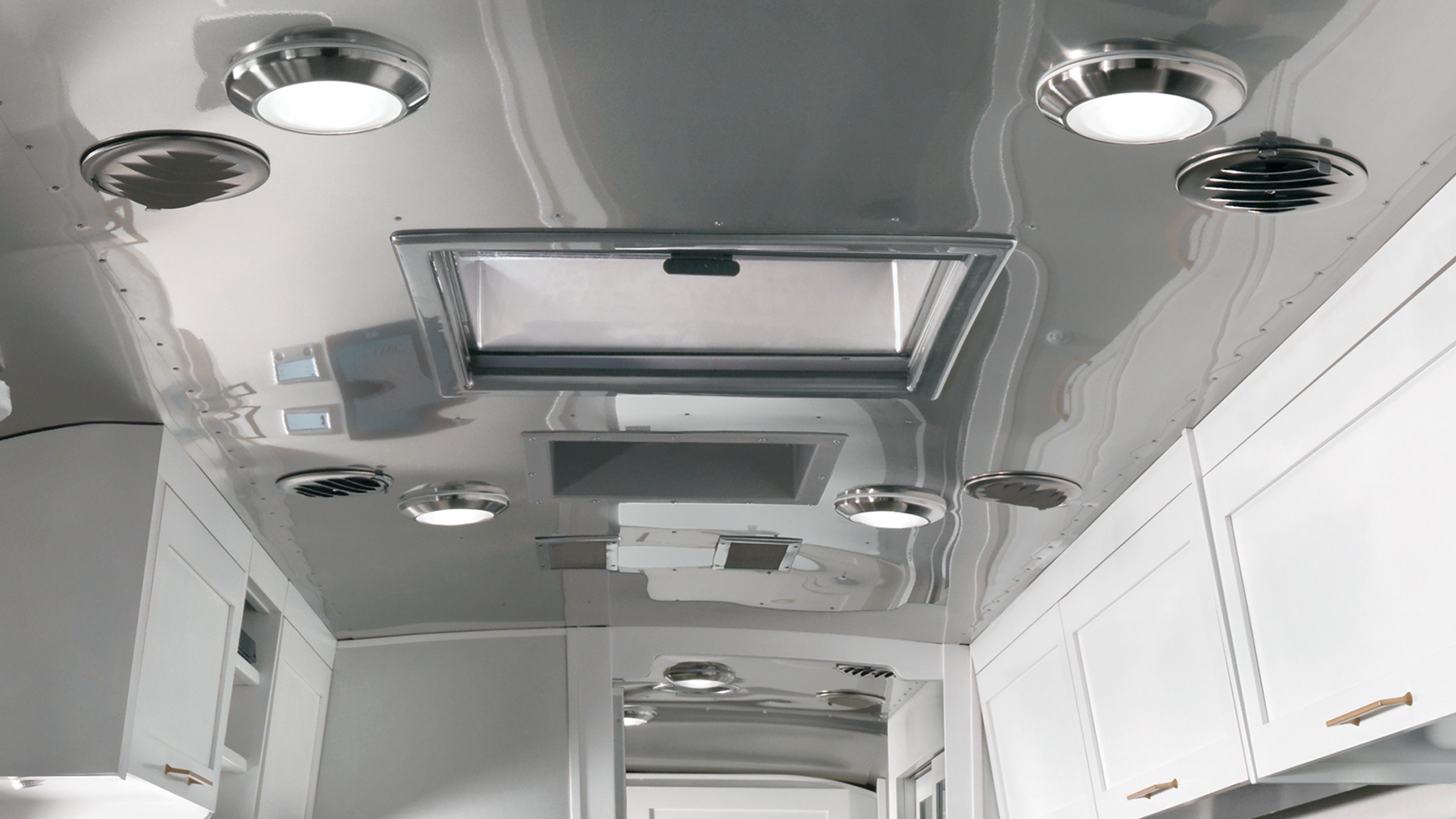 Interior view of a modern RV ceiling featuring round LED lights, circular air vents, a skylight, and white cabinetry below with brass handles. The surfaces are reflective and metallic.