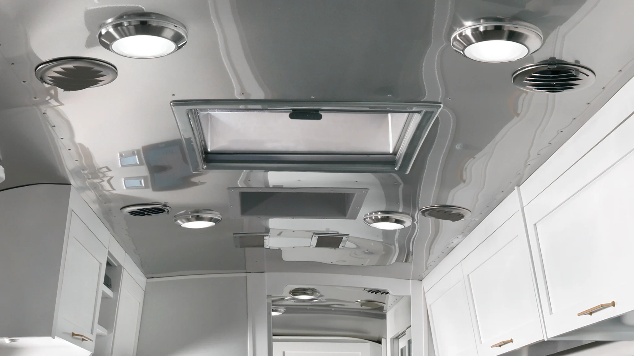 Interior view of a modern RV ceiling featuring round LED lights, circular air vents, a skylight, and white cabinetry below with brass handles. The surfaces are reflective and metallic.