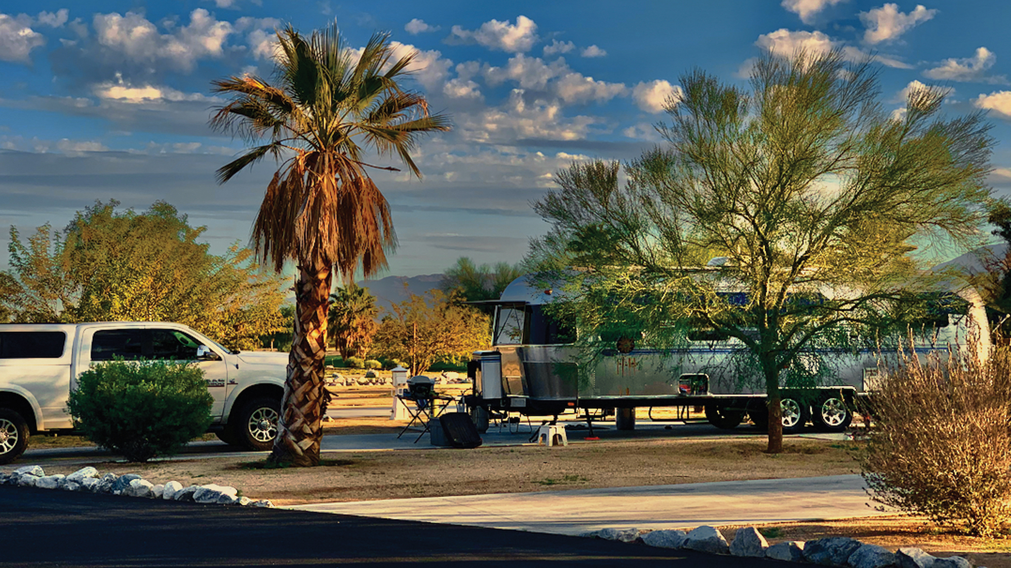 A white pickup truck and a silver trailer are parked at a desert campsite, surrounded by palm trees and shrubs under a blue sky with scattered clouds. Outdoor chairs and a dog are visible near the trailer.